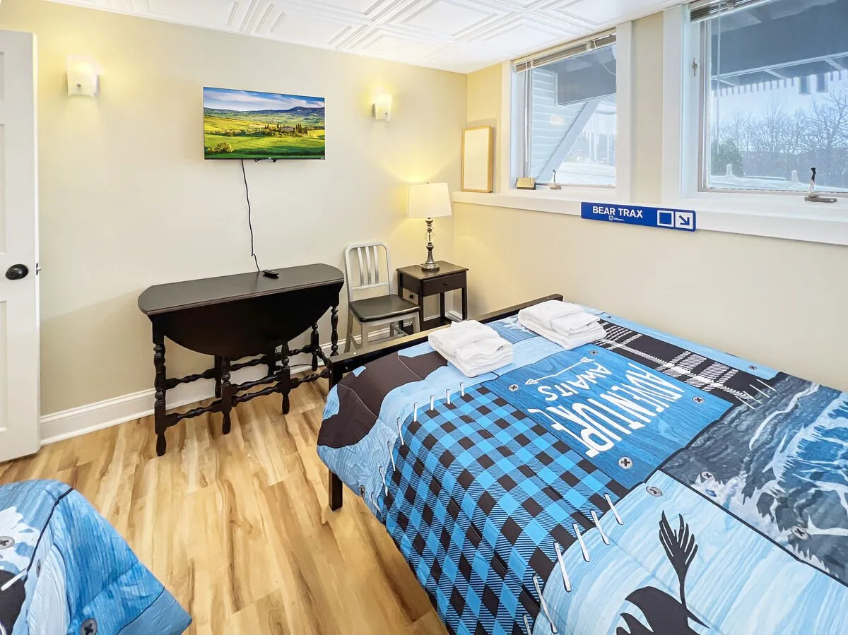 A cozy bedroom featuring bedding with a blue and black pattern, a landscape painting above the bed, a table lamp, and a small table with chairs. Natural light filters in, and the decor is inviting—perfect for relaxing after outdoor explorations or planning the next day’s adventure.