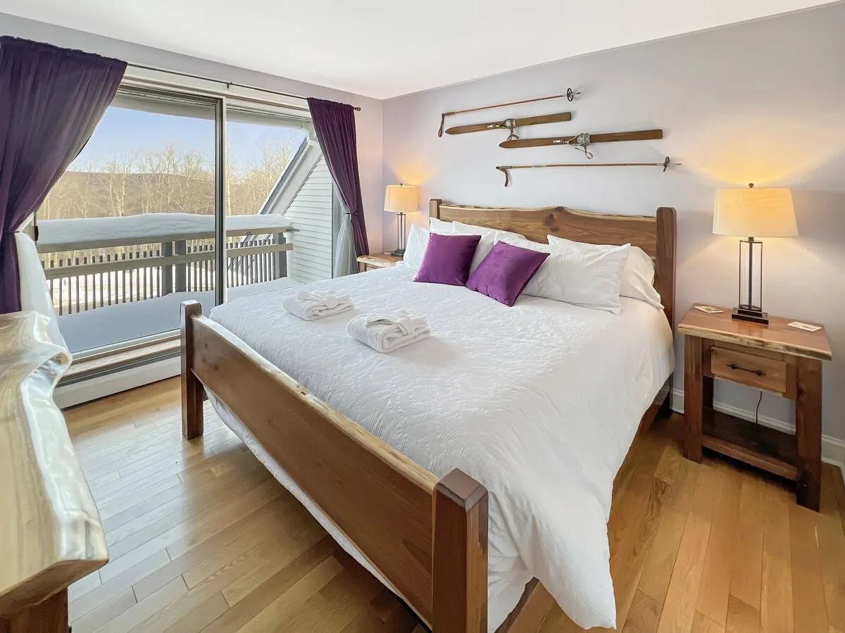 A comfortable bedroom on the main level, featuring a bed with crisp white linens and purple pillows, a large window showcasing snowy mountain views, and wooden accents that create a tranquil retreat.
