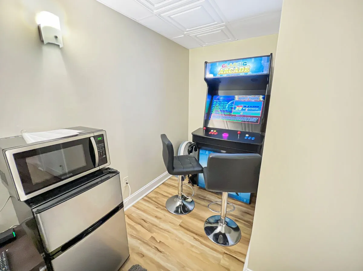 A lively recreation area featuring a classic arcade machine, bar stools, microwave, and refrigerator, creating a fun and informal space. The setup invites friendly competitions, snacks, and shared moments among guests of all ages.