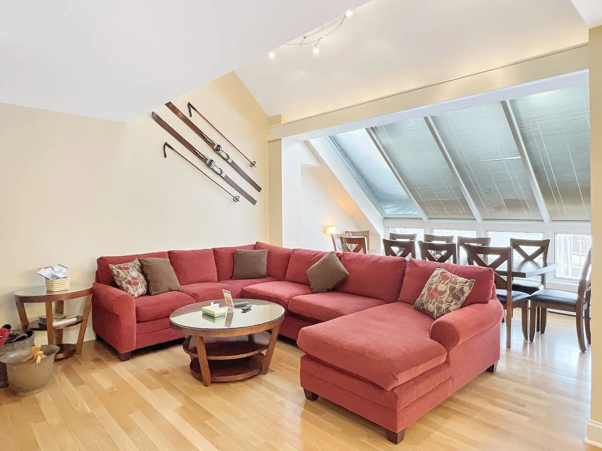 The image showcases a warm, inviting living space featuring a plush red sectional sofa surrounding a round coffee table, with a cozy dining area in the background framed by wooden chairs. Decorative ski equipment adorns the wall, blending rustic mountain charm with modern comfort—ideal for gatherings and après-ski relaxation.