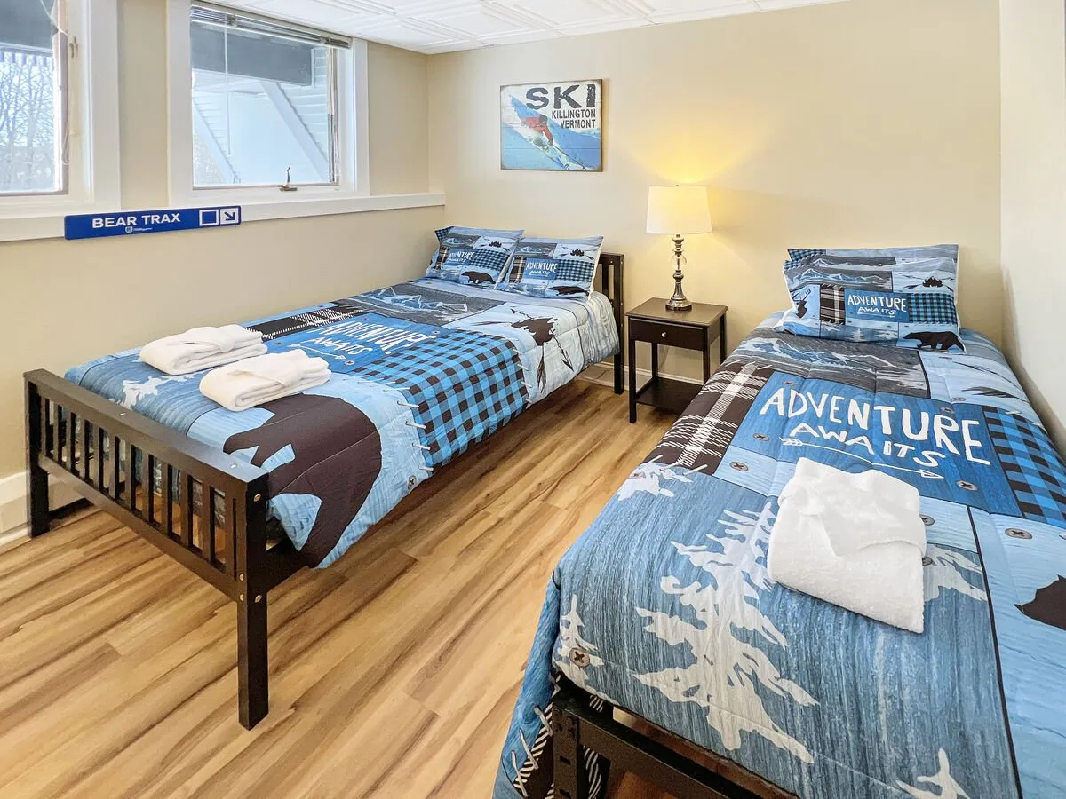 A cheerful second bedroom with two twin beds, colorful bedding, a lamp, wall art, and playful signage. The bright ambiance and mountain-themed decor create a welcoming, flexible space for families or friends.