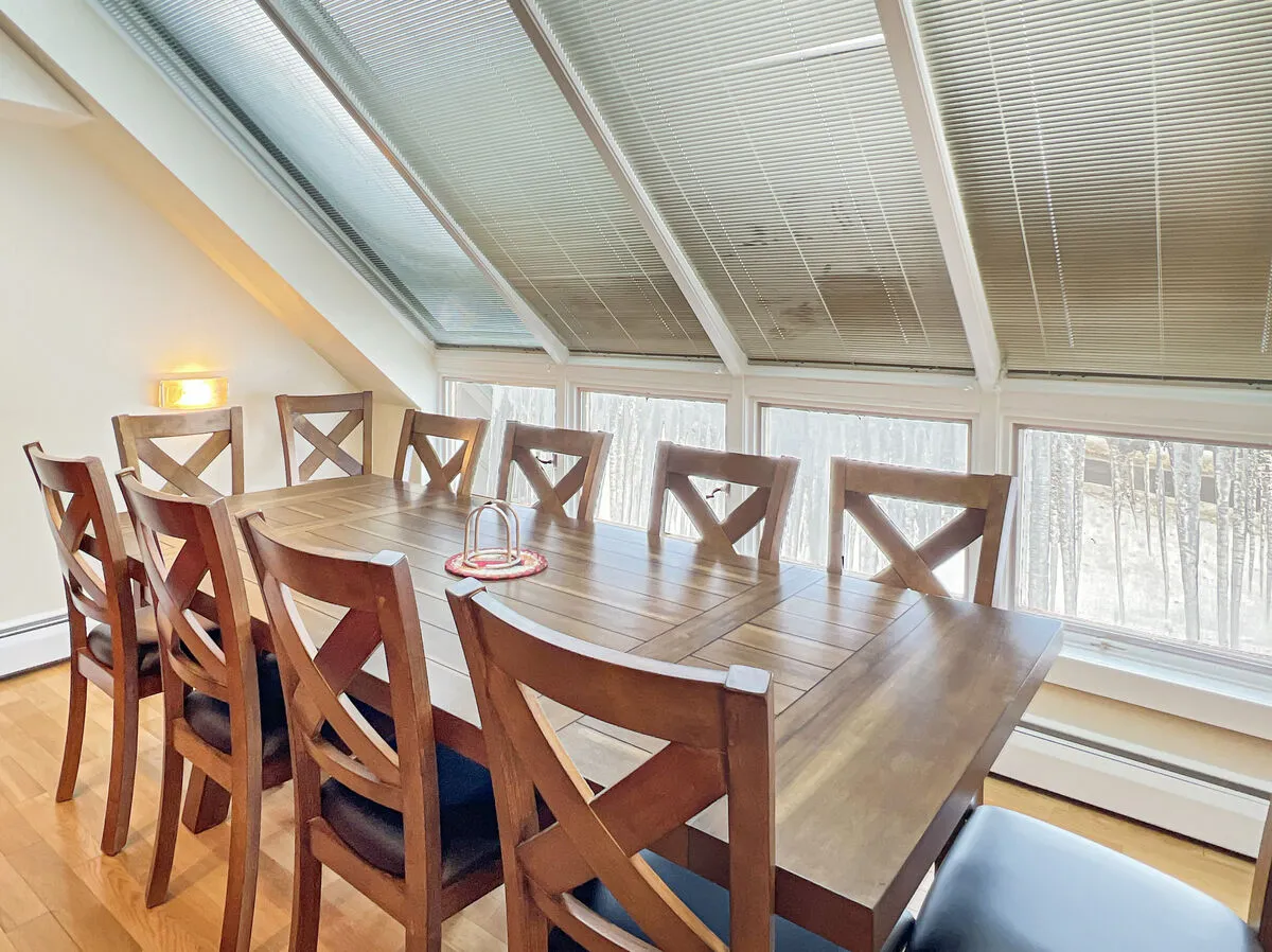 A spacious dining area with a large wooden table and chairs, illuminated by natural light streaming through expansive windows. The inviting setting is perfect for family meals, group celebrations, or casual conversation, capturing the spirit of communal mountain living.
