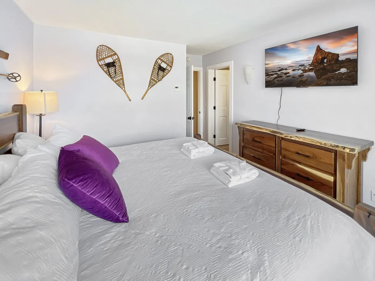 A spacious primary bedroom featuring a king-size bed with plush bedding, a decorative purple throw pillow, a wooden dresser, wall-mounted TV, and snowshoe wall art. The soft natural light filtering in adds to the serene, restful atmosphere.