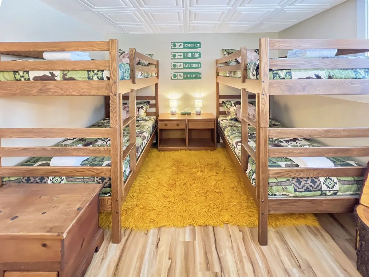 This image shows a playful kid-friendly bedroom with bunk beds dressed in bright bedding, a cheerful yellow rug, and nightstands with lamps. The decor includes wall signs, adding to the inviting and youthful atmosphere. The space feels designed for relaxation, play, and storytelling, ideal for families traveling together.