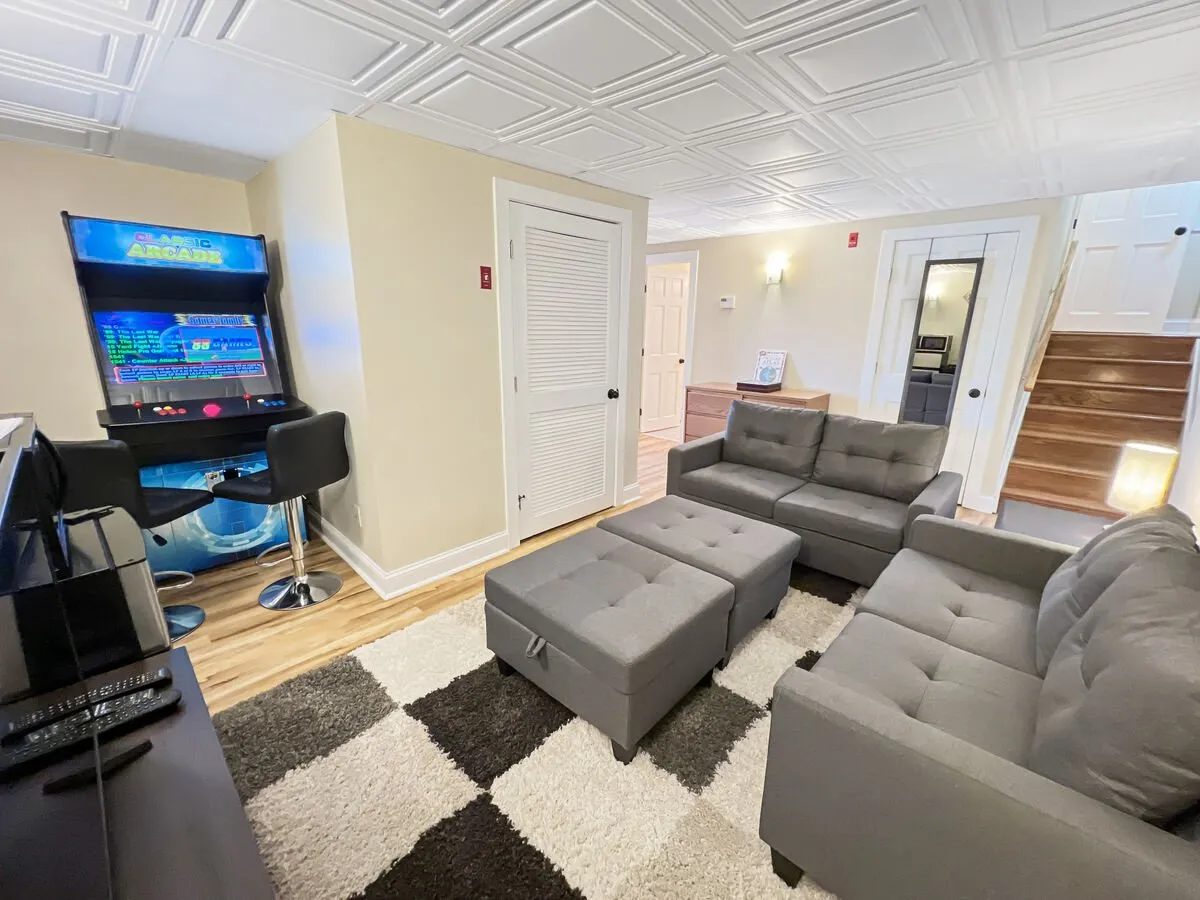 A playful and relaxed lounge area featuring an arcade machine, plush sofas, an ottoman, and a vibrant rug. The space is designed for fun game nights and relaxed socializing with friends or family.