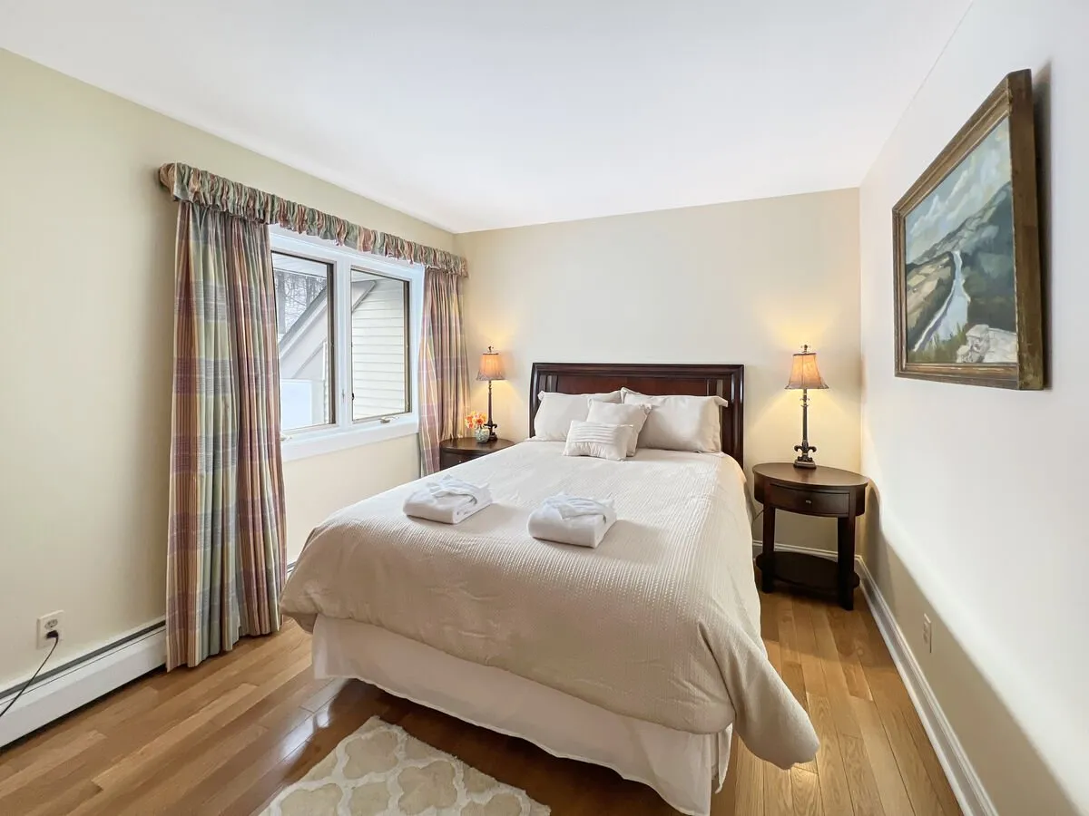 A serene bedroom featuring a cozy bed placed near a large window, a nightstand, and a painting. The window allows sunlight to stream in, illuminating the space and hinting at beautiful morning views. The overall mood is tranquil, suggesting a perfect start to the day for couples seeking relaxation.