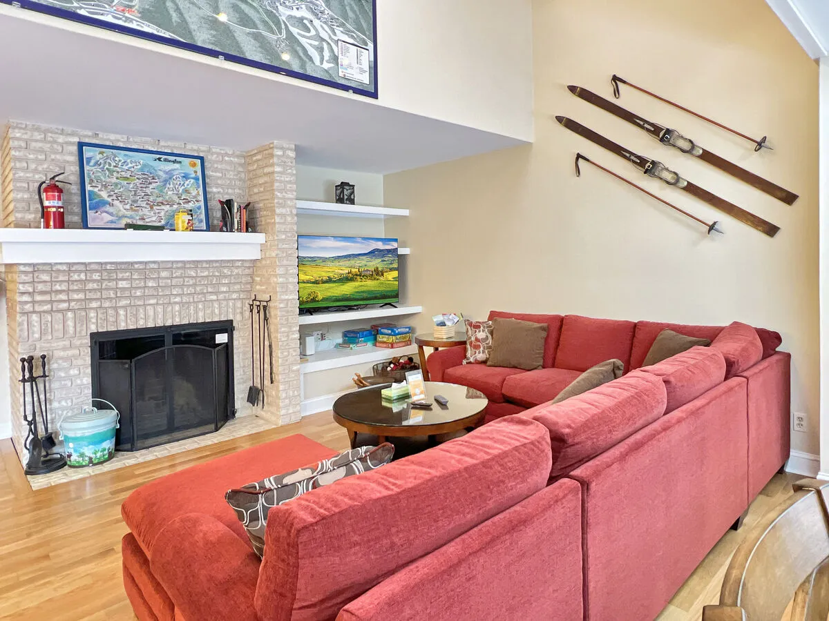 A warm and inviting living room featuring a red sectional sofa facing a wood-burning fireplace and a television. The space is decorated with ski-themed wall art and shelves filled with cozy decor. Large windows bring in ample natural light, creating a welcoming ambiance perfect for gatherings or relaxing after a day outside.